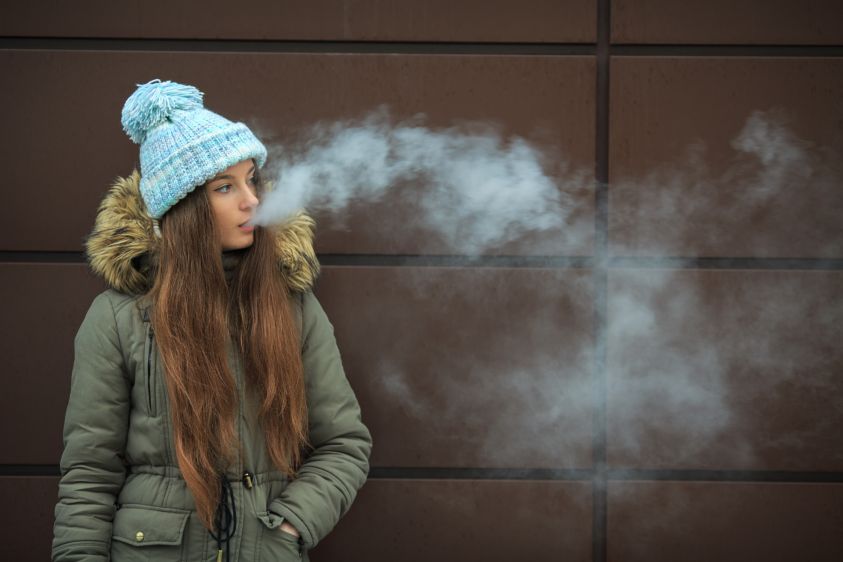 Vape teenager2e00 Young pretty white girl in blue cap is smoking an electronic cigarette opposite modern brown background on the street in the winter2e00 Bad habit2e00