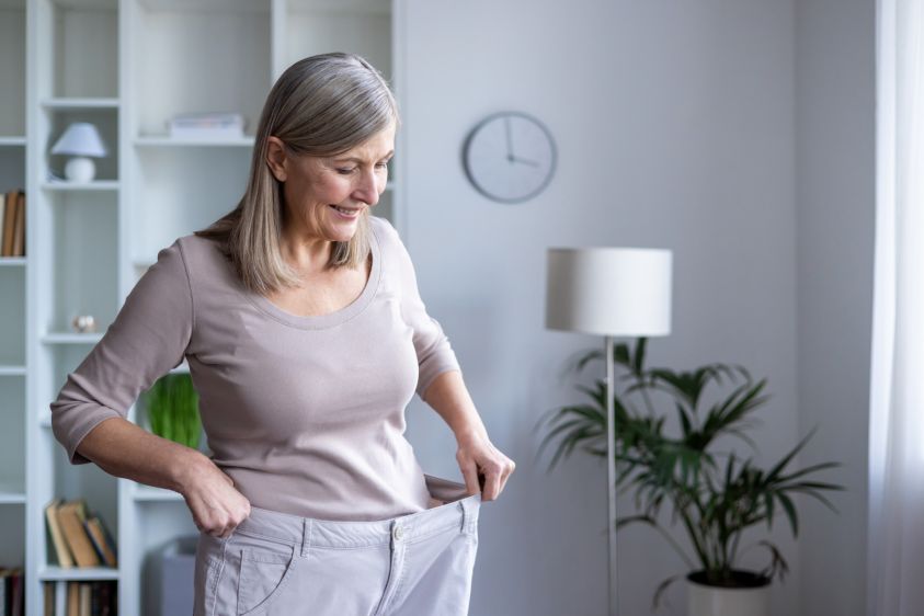 Smiling senior woman feels accomplished as she admires her weight loss progress in her large pants, standing in a cozy room, symbolizing healthy lifestyle and personal achievement2e00