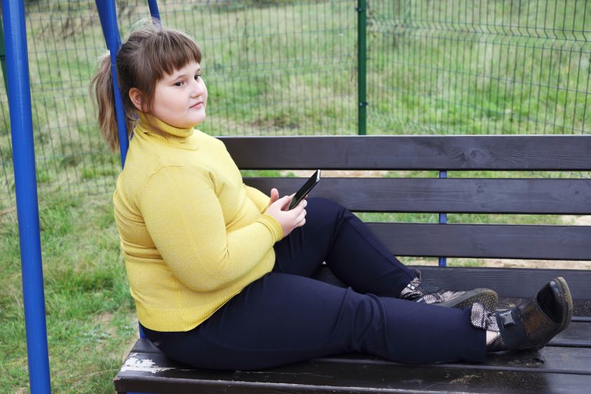 Plump thoughtful girl in yellow sweater sits alone on bench of swing outdoors, hold smartphone